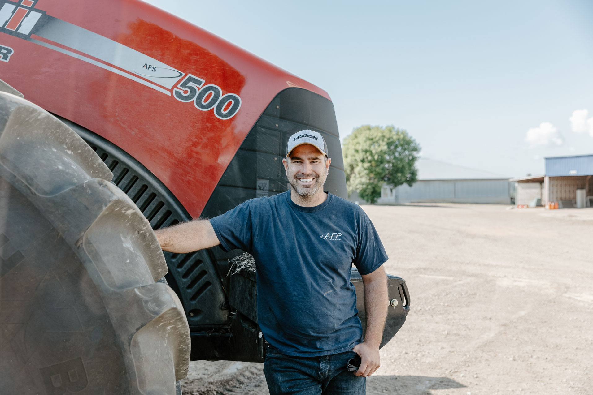 A man smiles, leaning against a tractor tire.