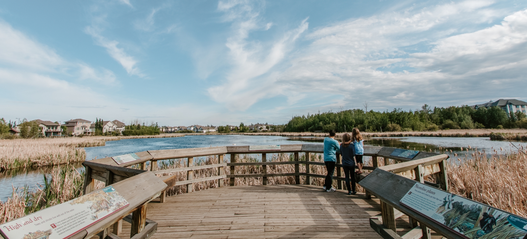 A family stands on a park boardwalk overlooking a pond.